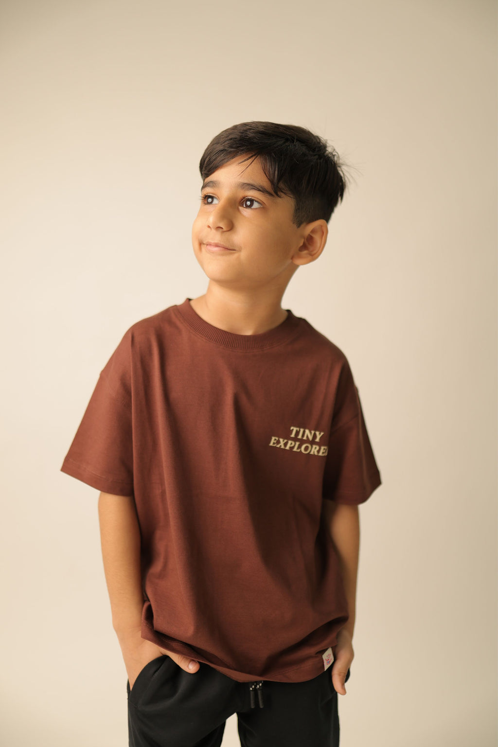 Brown kids' T-shirt on model with small front “TINY EXPLORER” print, short sleeves
