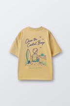 Sand yellow graphic tee with surf-kid illustration and 'Chase the Coastal Breeze' text.