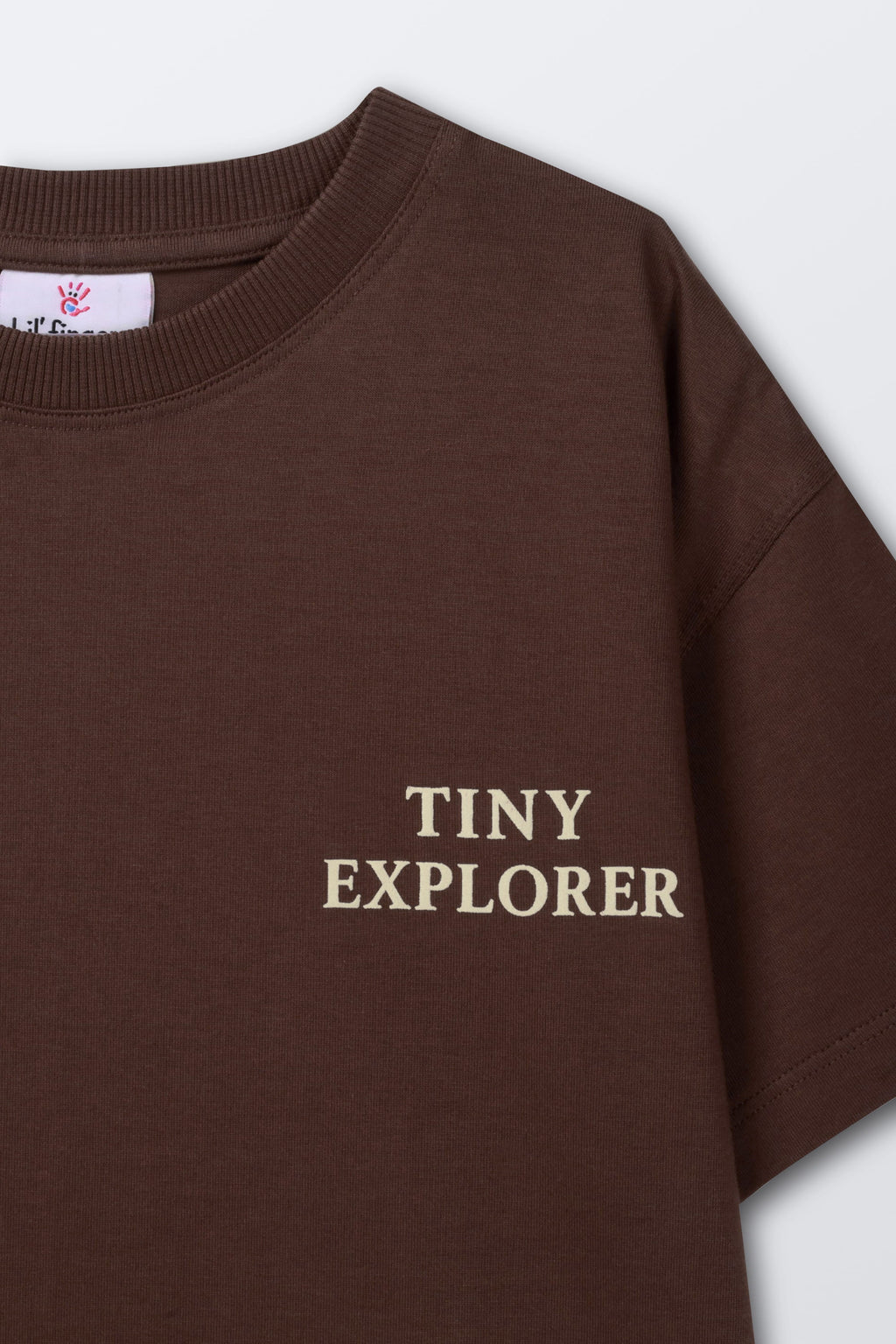 Brown kids' T-shirt side close-up showing “TINY EXPLORER” front text
