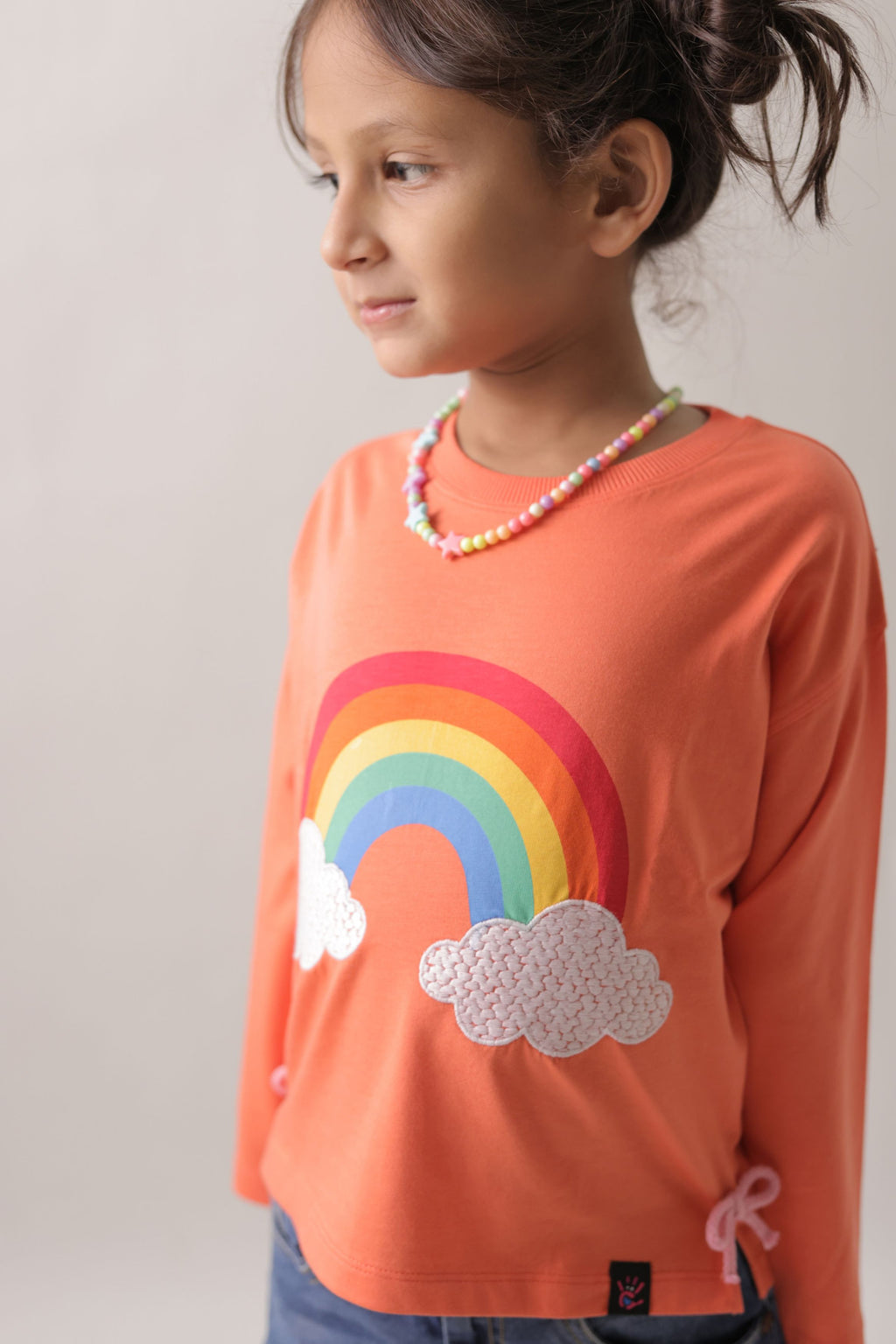 Girl wearing coral T-shirt front with rainbow, embroidered clouds, and bow hem ties