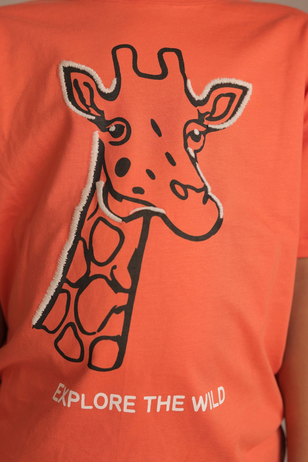 Orange comfort tee close-up with embroidered giraffe and 'EXPLORE THE WILD' text