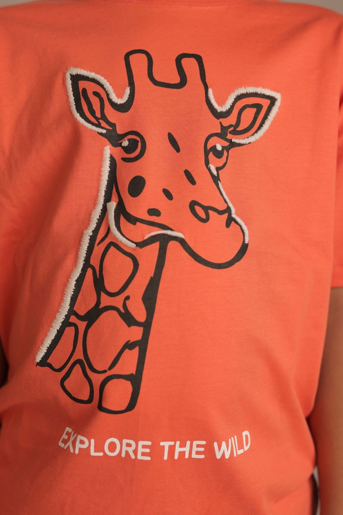 Orange comfort tee close-up with embroidered giraffe and 'EXPLORE THE WILD' text