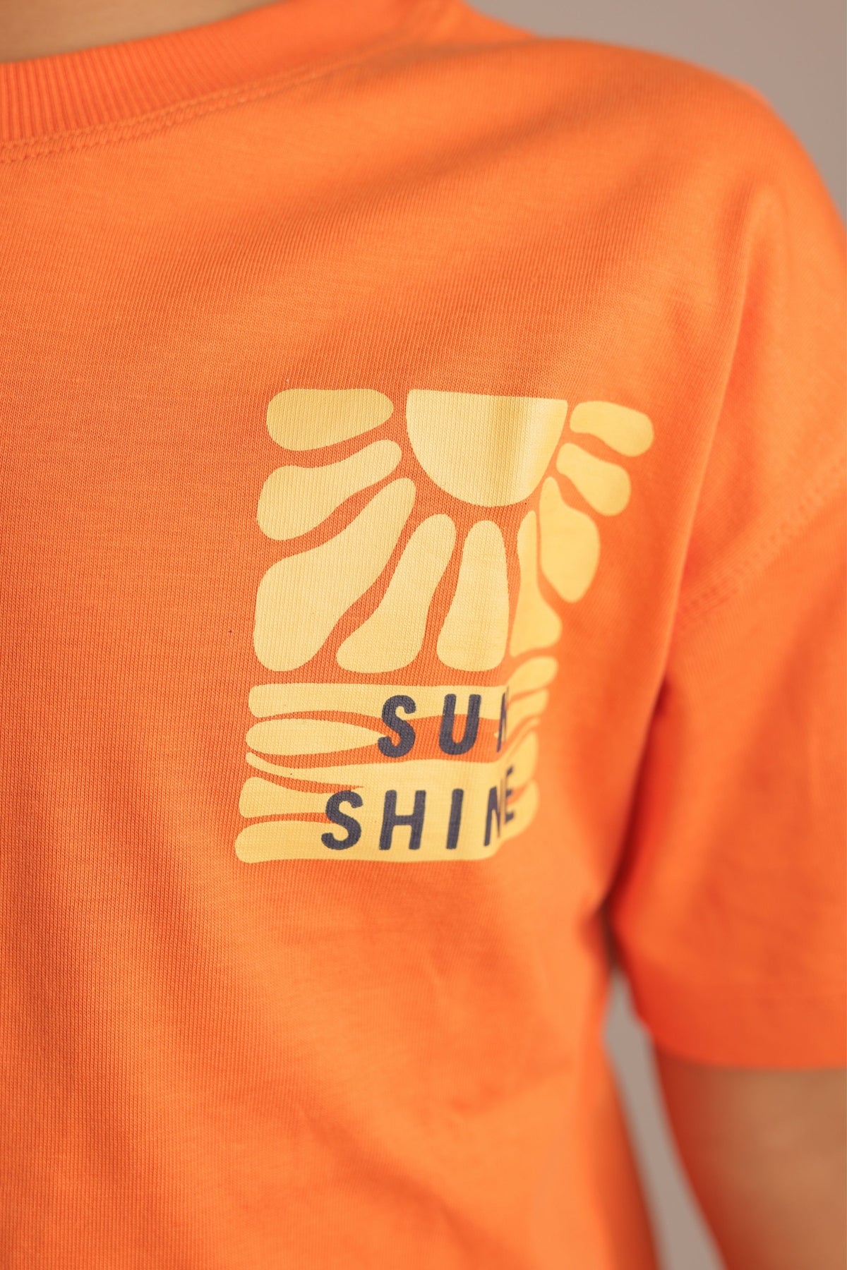 Orange kids' tee close-up: 'Sun Shine' sun icon graphic on back